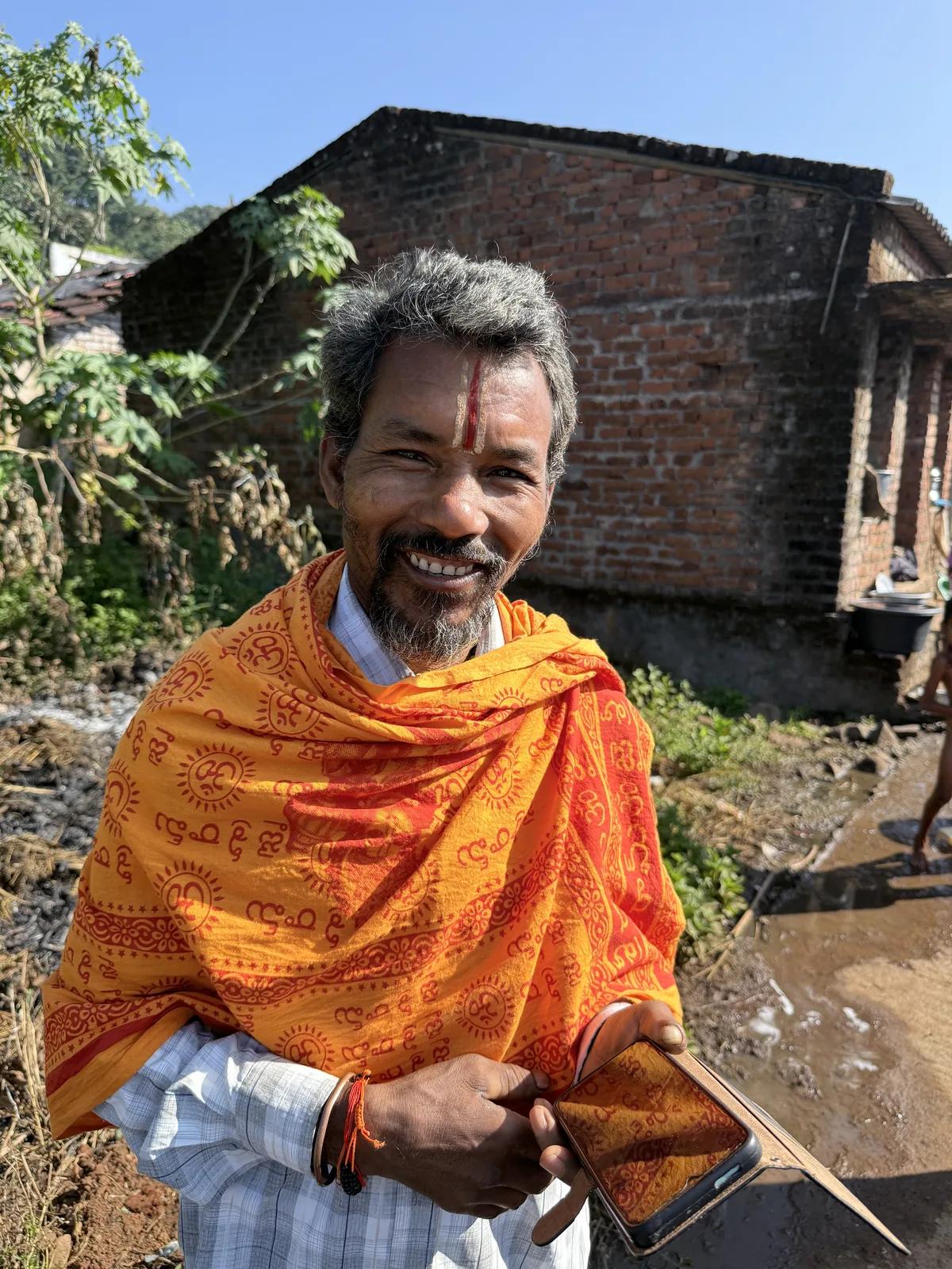 A smiling man in an orange shawl stands outside a brick home in Logilli.