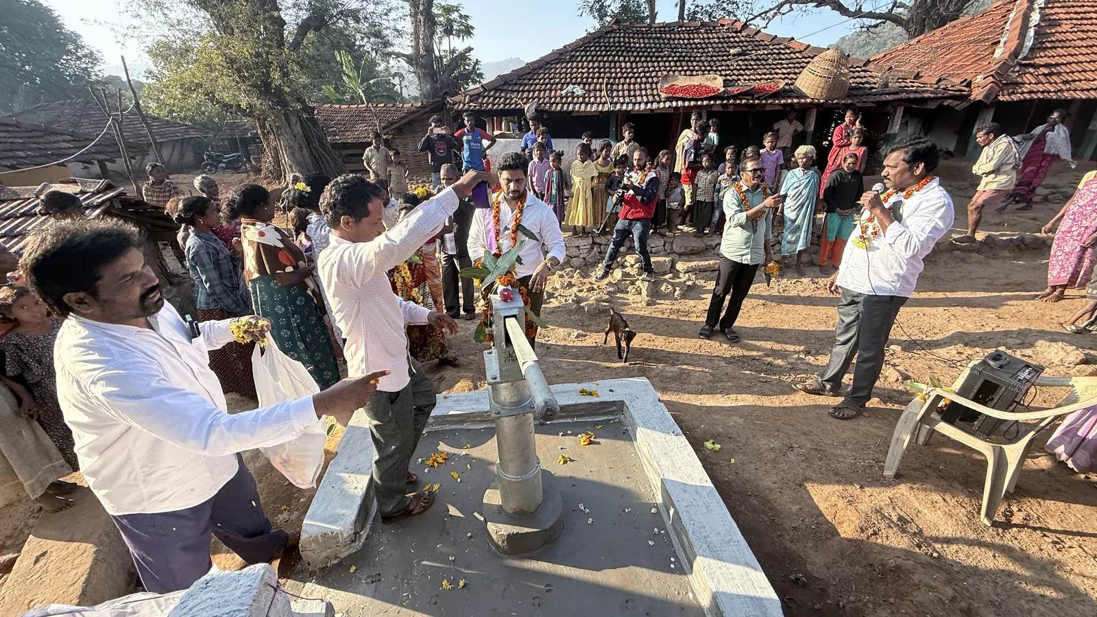 Men wearing flower garlands stand at a new hand pump decorated with flowers, as villagers and children gather among tiled-roof homes.