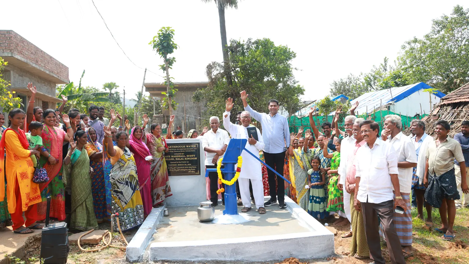 Community members raise their hands around a newly opened blue hand pump during a Breakthrough Water well dedication.