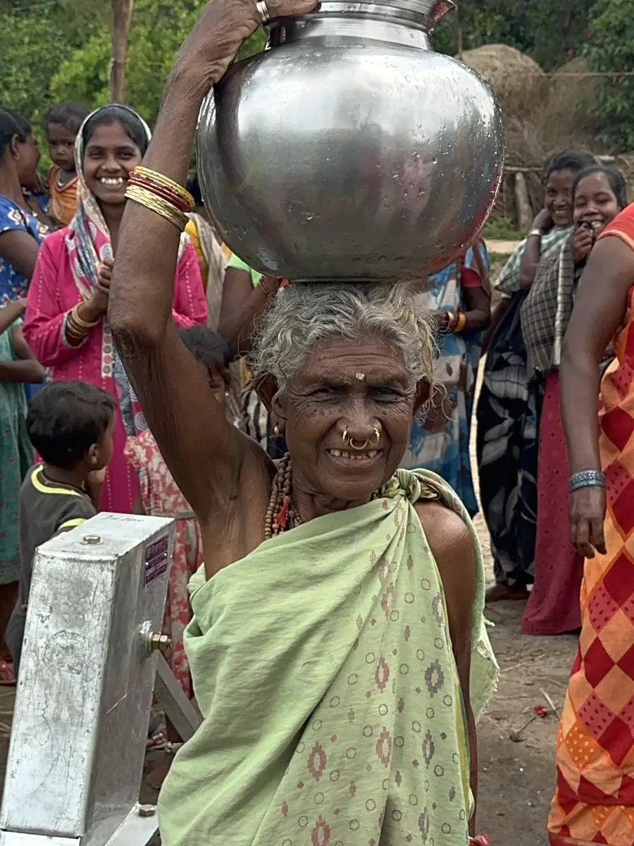 A woman carries a metal water vessel through a rural village.