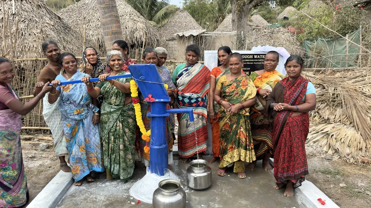 Women gather with metal vessels around a blue village hand pump flowing with clean water.