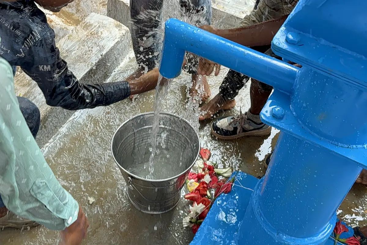 Clean water flows from a blue hand pump into a bucket held below.