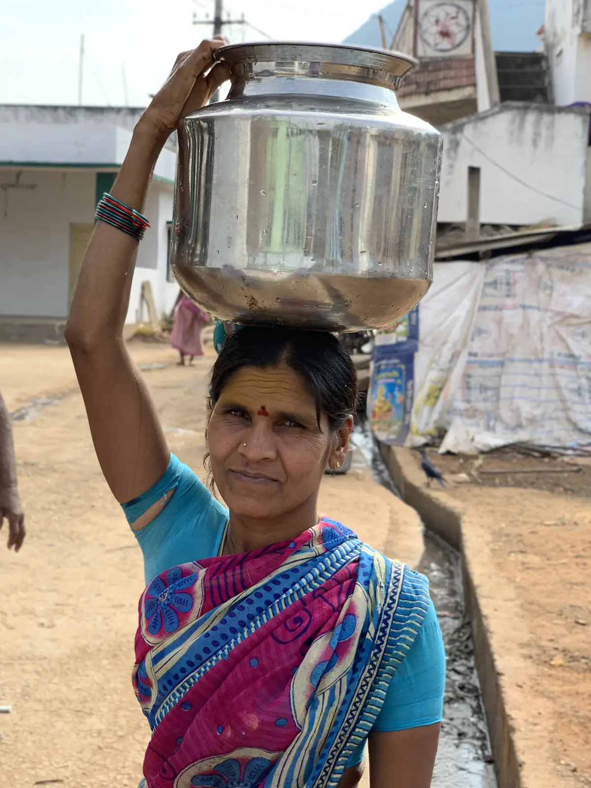 A woman balances a metal water vessel on her head in a rural village.