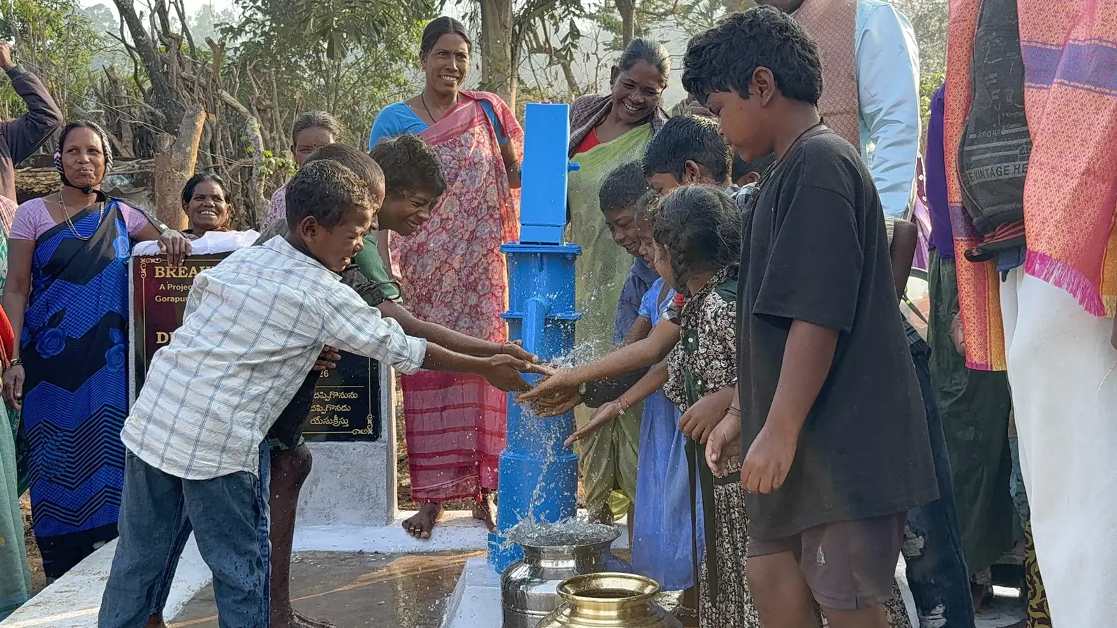 Children and women gather joyfully around a blue village hand pump flowing with clean water.