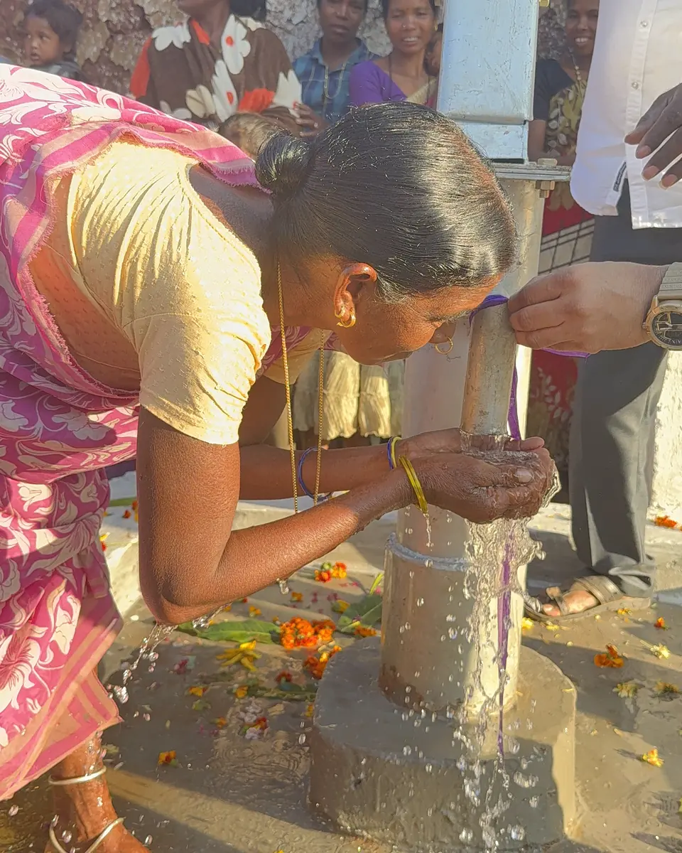 A woman drinks directly from a village hand pump as neighbors gather nearby.