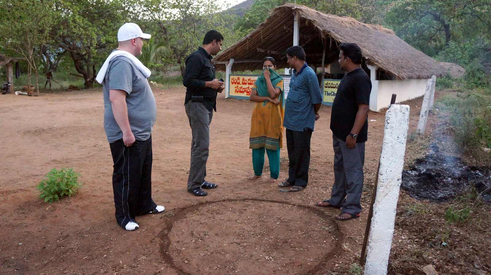 A small group stands beside a circle marked on the ground for drilling, next to a thatched building in Makkuva.