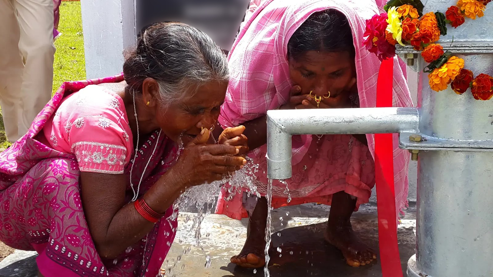 Two women drink water beside a village hand pump.